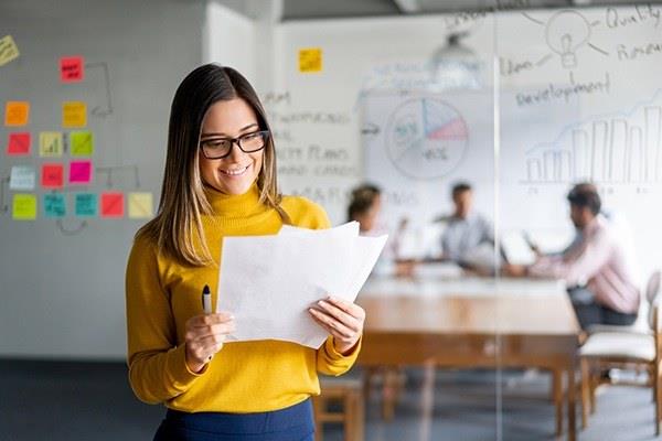 person holding papers standing in office with meeting in background