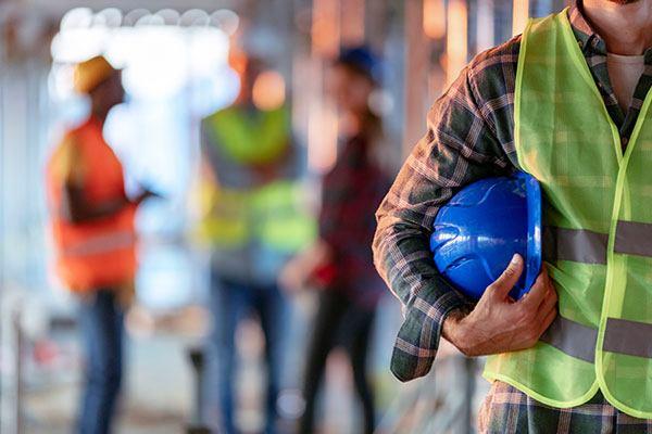 person in flannel and safety vest holding a hardhat with other safety workers in background