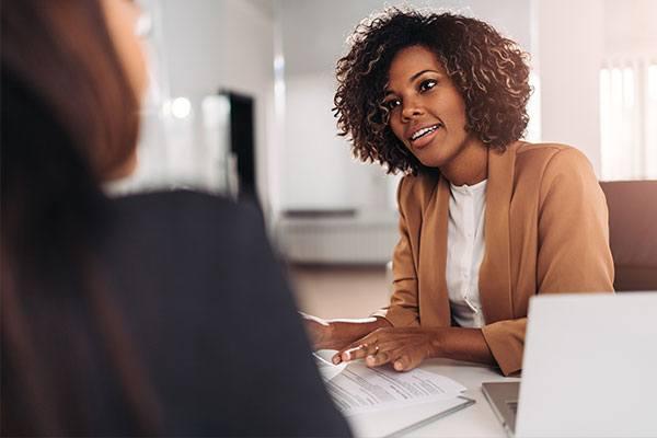 woman talking with someone across a desk with laptop and book