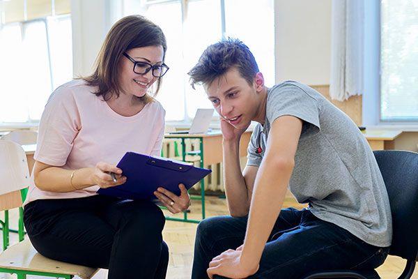 smiling teacher in classroom with clipboard sitting next to smiling student