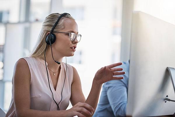 woman in glasses wearing headset talking and looking at computer monitor