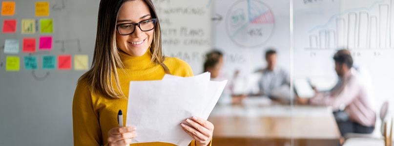 woman looking at papers standing in front of wall of sticky notes with table of staff meeting in conference room