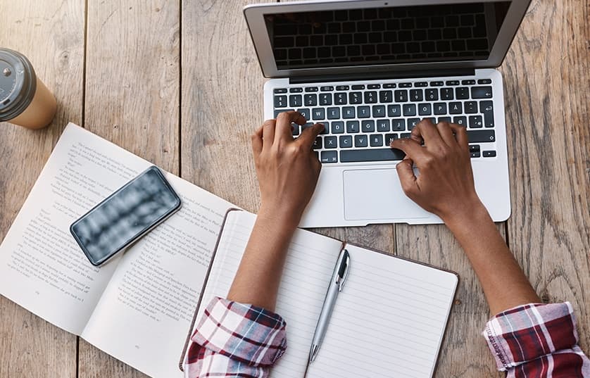 student working on a laptop with a book, notebook, coffee and phone